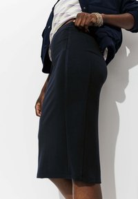 Personne portant une jupe noire à mi-longueur, un haut rayé et une veste bleu marine, montrant un poignet orné de bracelets près de la taille.