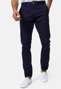 Navy blue cotton chinos with a tailored fit, featuring front pockets, a buttoned waistband, and a smooth texture. Pants paired with white slip-on shoes.