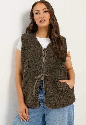 Femme portant un gilet sans manches marron texturé noué devant par-dessus une chemise blanche et un jean bleu, main dans la poche du gilet.