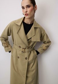 Bež plašč z pasom, dvojnimi gumbi in čezmernimi ovratniki. Ima kovinske detajle na manšetah in žepih.