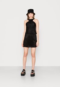 Vestido preto plissado com decote halter e design em camadas. Usado com um chapéu preto de balde e sandálias de plataforma, criando um look coordenado.
