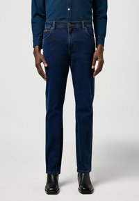 Dark blue denim jeans with a straight fit, five pockets, and a button closure. Paired with black ankle boots and a dark button-up shirt.