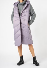 Ljuslila, ärmlös puffer väst med huva, diagonal sömnad och tryckknappar. Bärs över en grå tröja med vida ärmar.