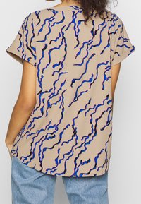 Beige top s krátkými rukávy, zdobený vlnkovaným vzorem v černé, modré a červené barvě, vyrobený z lehkého materiálu, s volným střihem.