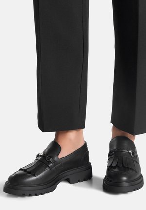 Schwarze Leder-Loafers mit dicker Sohle, verziert mit Fransen und metallischen Akzenten. Kombiniert mit maßgeschneiderten schwarzen Hosen.