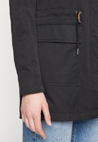 Svart jacka med strukturerad tyg, synliga sömmar och en framficka. Hand som vilar på kanten av jackan.