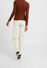 Personne marchant de dos, portant un haut à manches longues côtelé de couleur rouille, un pantalon en velours côtelé blanc et des baskets noires et blanches sur un fond uni.