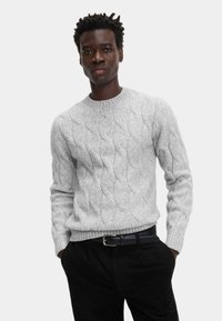 Grau gestrickter Pullover mit einem Zopf-Muster und geripptem Kragen. Über einer schwarzen Hose getragen, mit einem schwarzen Gürtel, der eine silberne Schnalle hat.