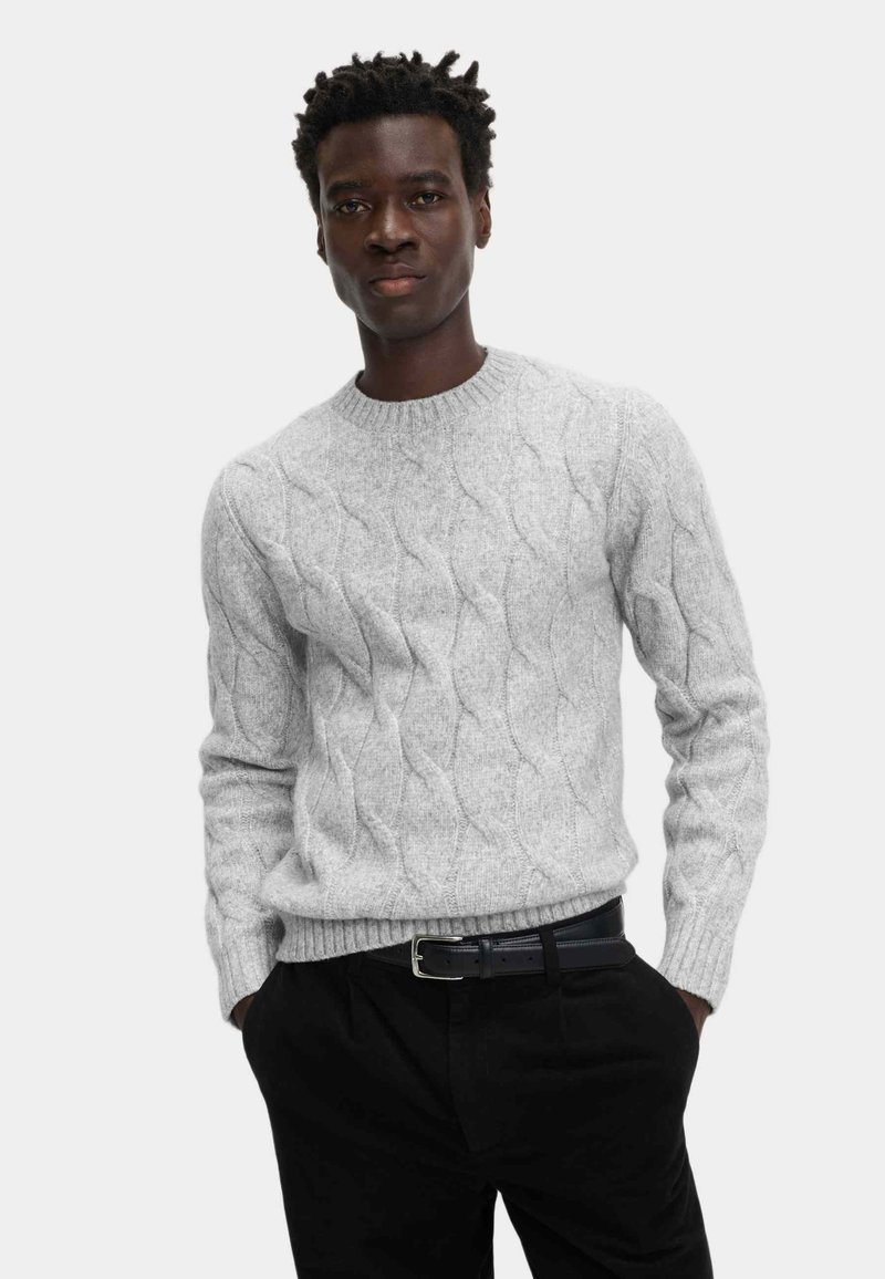 Grau gestrickter Pullover mit einem Zopf-Muster und geripptem Kragen. Über einer schwarzen Hose getragen, mit einem schwarzen Gürtel, der eine silberne Schnalle hat.