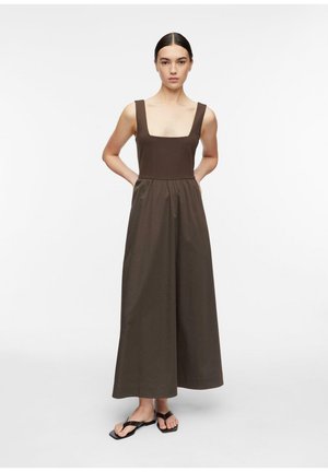 Kobieta stoi z rękami za plecami, ubrana w brązową sukienkę maxi bez rękawów i czarne sandały na jednolitym białym tle.