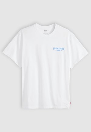 Camiseta blanca de manga corta con un pequeño texto azul que dice "LIVING LEGEND" impreso en el pecho izquierdo sobre un fondo gris liso.