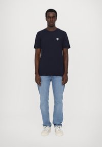 Tamsiai mėlyna marškinė su apvaliu iškirpimu ir trumpomis rankovėmis, su mažuoju sidabro logo kairėje krūtinės pusėje. Dėvimi su šviesiai mėlynomis džinsomis.