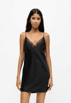 Femme aux longs cheveux noirs portant une robe nuisette noire sans manches avec des bordures en dentelle, debout devant un fond clair uni.