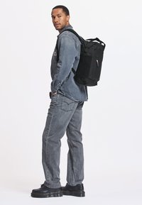 Uno zaino nero presenta una forma rettangolare con spallacci imbottiti, tasca esterna con zip e un dettaglio di portachiavi colorato. Indossato da una persona con una giacca di jeans.