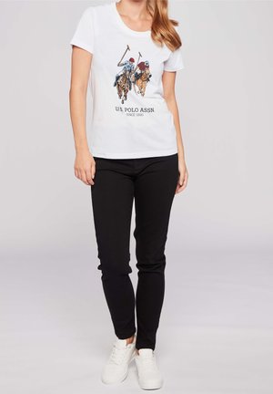 Bílé tričko s krátkým rukávem, na kterém je barevný obrázek dvou jezdců na koních při polo. Text zní "U.S. POLO ASSN. SINCE 1890." Černé kalhoty a bílé tenisky.