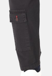 Gamba di pantaloni cargo neri con una tasca con bottone sopra la caviglia, indossata con calze scure e sneakers bianche su uno sfondo bianco.