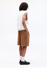 Hvid ærmeløs sweatshirt og brune shorts, ensfarvet tekstur, sidetasker, parret med sorte sko og hvide ankelstrømper, stående i profil.