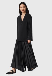 Vrouw in een zwart sweater met diepe V-hals, geplooide jumpsuit met wijde benen en zwarte puntige flats met enkelbandjes, staand tegen een lichte achtergrond.