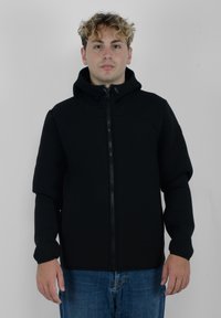 Giacca nera con zip e cappuccio, caratterizzata da una texture liscia e un design aderente. I polsini sono aderenti, in abbinamento a jeans blu denim casual.