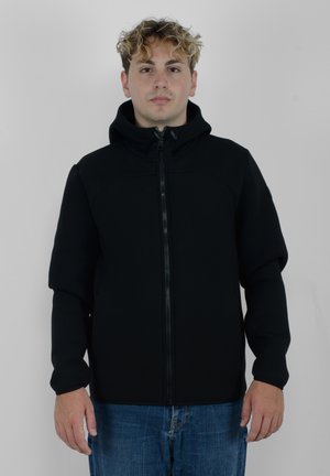 Giacca nera con zip e cappuccio, caratterizzata da una texture liscia e un design aderente. I polsini sono aderenti, in abbinamento a jeans blu denim casual.