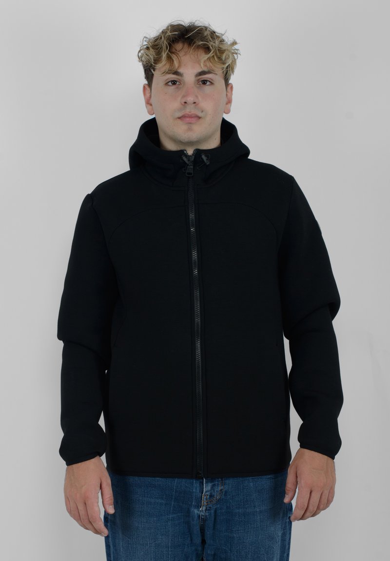 Giacca nera con zip e cappuccio, caratterizzata da una texture liscia e un design aderente. I polsini sono aderenti, in abbinamento a jeans blu denim casual.