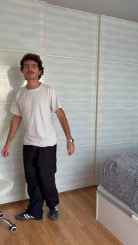 Jeune homme portant un t-shirt blanc et un pantalon noir se tient dans une pièce avec un sol en bois, un mur à panneaux blancs ondulés, une chaise de bureau et un lit avec une couverture grise.