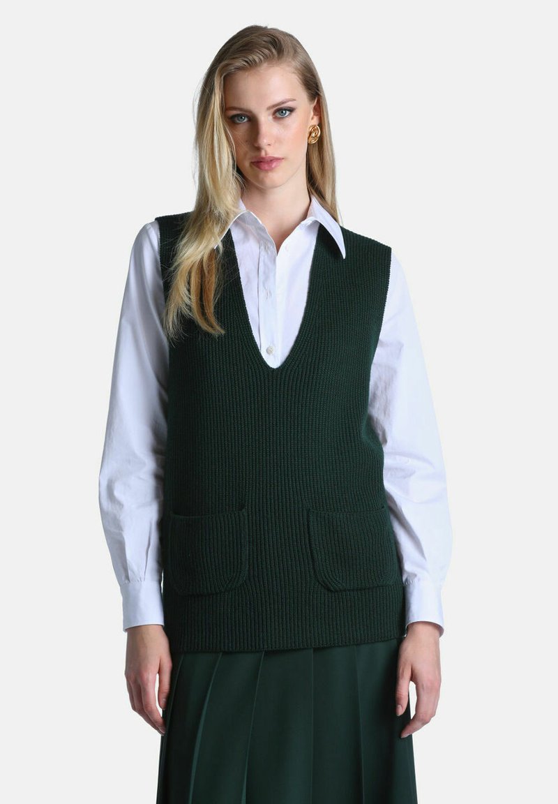 Femme portant un gilet en maille vert foncé avec des poches sur une chemise blanche boutonnée et une jupe plissée verte assortie, debout devant un fond blanc.