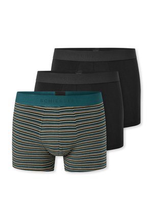 Schlanker Mann mit kurzen lockigen Haaren, der grün-braun gestreifte Boxershorts trägt, steht vor weißem Hintergrund.