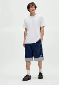 Mladý muž stojí s rukami v vreckách, oblečený v bielom tričku, tmavomodrých a šedých basketbalových šortkách, bielych ponožkách a tmavohnedých topánkach.