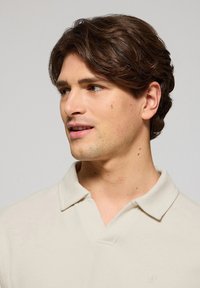 Polo shirt beige chiaro con un tessuto testurizzato e un collo classico. Presenta un logo sottile sul lato sinistro del petto. Vestibilità liscia e strutturata.