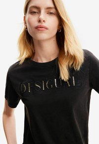 Camiseta de algodón negra con letras recortadas "DESIGUAL" adornadas con tachuelas metálicas. Manga corta y escote redondo.
