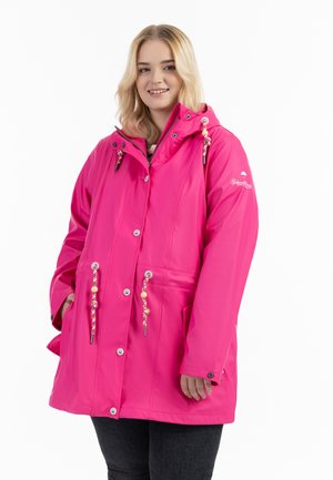 Junge Frau mit blondem Haar, die einen leuchtend pinken Regenmantel mit Kapuze, vorderen Knöpfen und bunten Kordeln trägt, steht mit den Händen in den Taschen.
