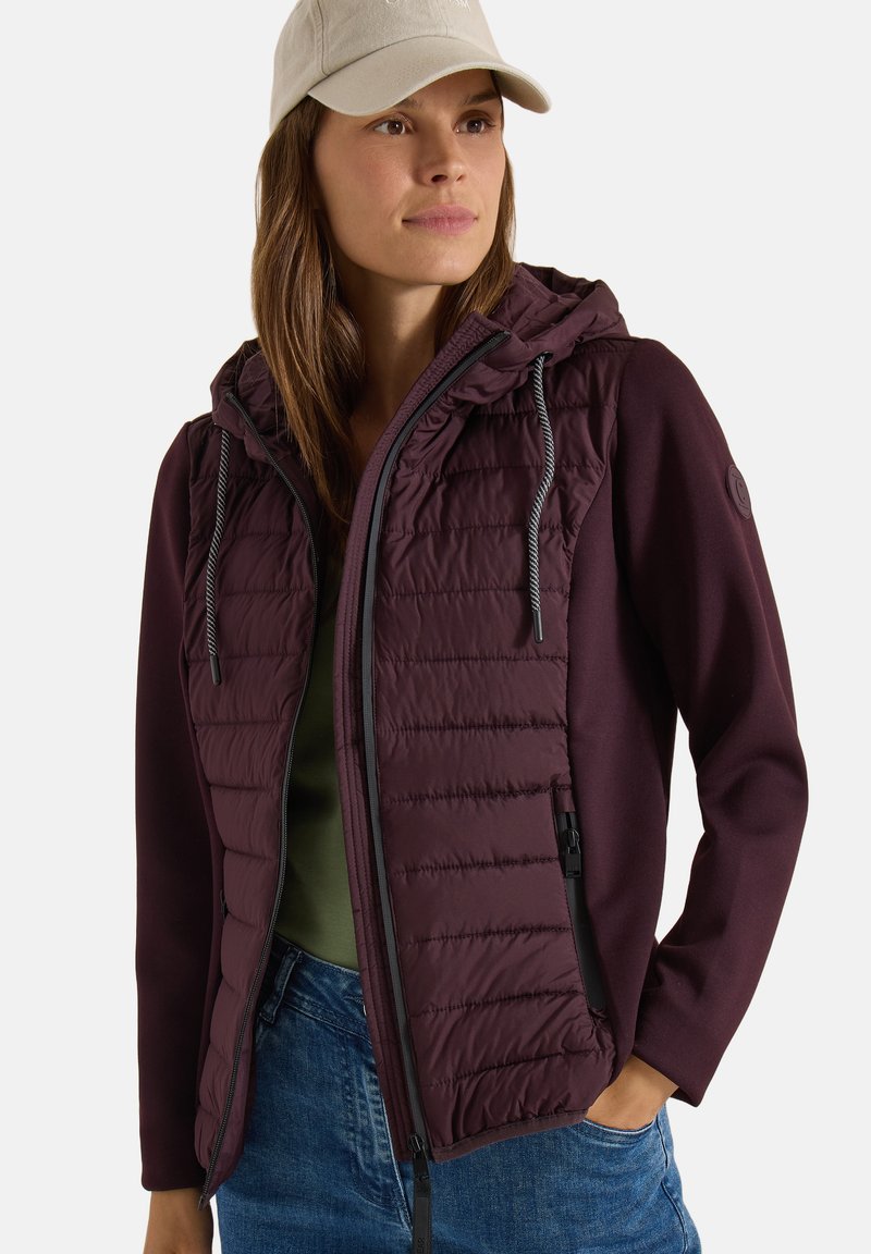 Femme portant une veste matelassée bordeaux avec une capuche, une chemise verte, un jean bleu et une casquette de baseball beige, posant avec une main dans la poche.