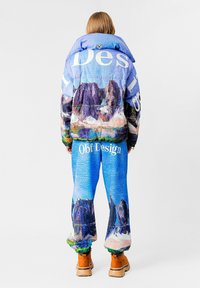 Una chaqueta y pantalones acolchados en bloques de color presentan un estampado de paisaje montañoso en azul, púrpura y verde, con texto de marca en blanco. Se pueden ver calzado marrón.