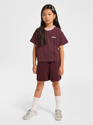 Ung pige med langt sort hår, iført mørkerød t-shirt med striber og shorts, hvide sokker og sølvfarvede sneakers, stående mod en hvid baggrund.