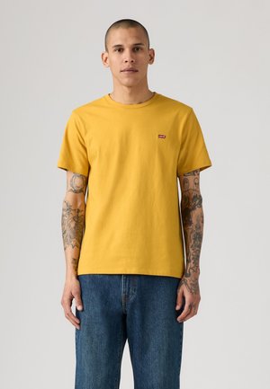 Homme aux cheveux courts et aux bras tatoués porte un t-shirt Levi's jaune moutarde et un jean bleu, debout devant un fond uni.