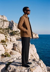 Uomo con giacca trapuntata marrone, pantaloni neri e occhiali da sole in piedi su una scogliera rocciosa con vista sul mare blu sotto un cielo limpido.