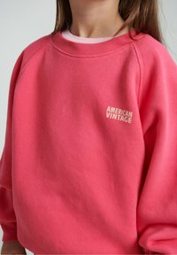 Jasnoróżowa bluza z okrągłym dekoltem, rękawami raglanowymi oraz białym logo "AMERICAN VINTAGE" na przodzie. Miękki, fakturowany materiał.