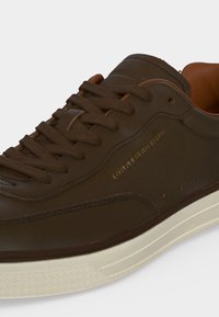 Καφέ δερμάτινο αθλητικό παπούτσι Tommy Hilfiger με σκούρα καφέ κορδόνια και λευκή λαστιχένια σόλα, με πλευρικό λογότυπο και λεπτομερείς ραφές.