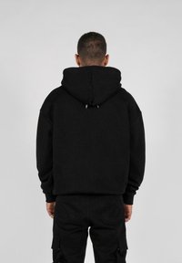 Schwarzer Kapuzenpullover mit lockerer Passform. Verfügt über eine Kordelzugkapuze und zwei auffällige weiße Akzente am Kragen. Kombiniert mit dunklen Hosen.