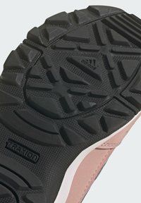 Czarna podeszwa buta sportowego z głębokimi wzorami trakcjami, w tym piramidalnymi wypustkami. Bok charakteryzuje się gładkim różowym i białym elementem wzorniczym.