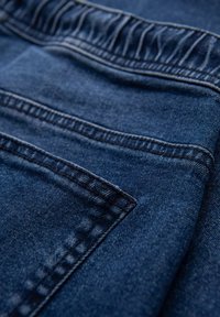 Tela de mezclilla azul oscuro con una textura suave, que presenta un bolsillo visible y costuras clásicas a lo largo de las uniones.