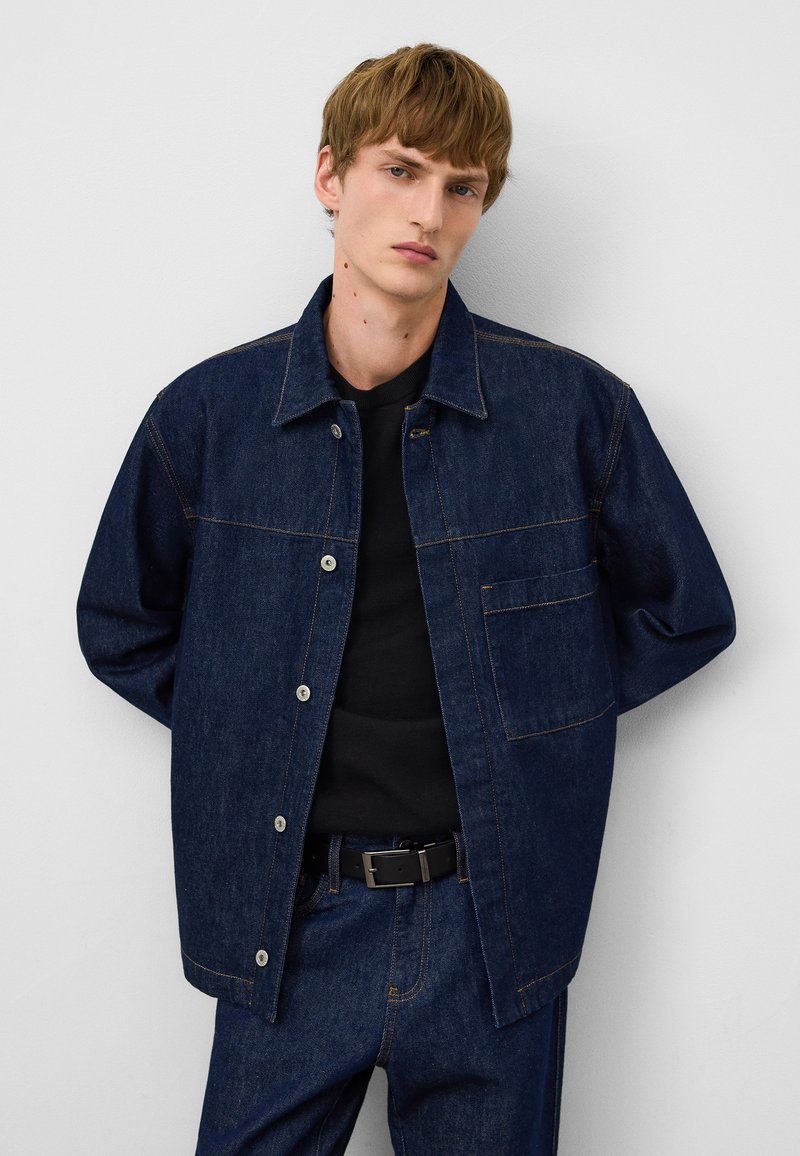 Denimjacka i mörkblått med knappstängning, två sidofickor och kontrasterande sömmar. Bärs över en svart topp och matchande byxor.