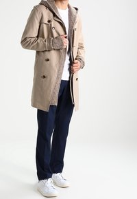 Beige vattentålig kappa med huva, texturerat grått foder, dubbla bronsknappar, bärs över en vit skjorta och marinblå byxor, ihop med vita sneakers.