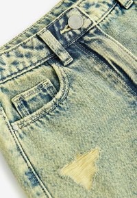 Gros plan sur un jean bleu délavé à l’acide et usé, montrant la poche avant, le passant de ceinture, le bouton en métal et une grande déchirure près de la poche.