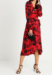 Röd blommig omlottklänning med lång ärm, med svarta blommönster. Klänningen har en sidoklyfta och är kombinerad med svarta klackar och en kuvertväska.