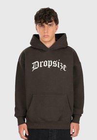 Junger Mann, der einen dunkelbraunen Kapuzenpullover mit "Dropsize" in weißer Gotikschrift trägt und vor einem schlichten hellgrauen Hintergrund steht.
