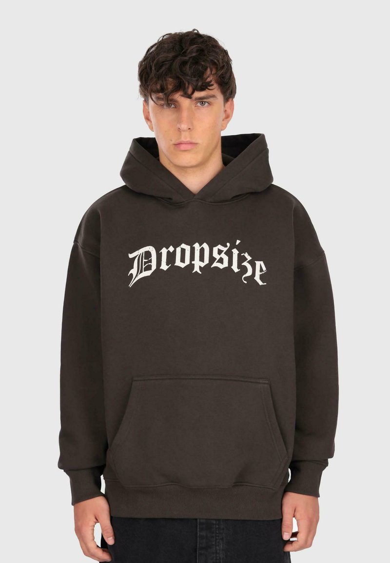 Junger Mann, der einen dunkelbraunen Kapuzenpullover mit "Dropsize" in weißer Gotikschrift trägt und vor einem schlichten hellgrauen Hintergrund steht.