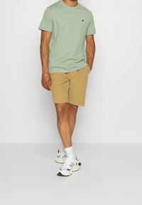 T-shirt en coton vert clair avec un petit logo noir, associé à un short beige et des baskets blanches, mettant en avant un style simple et décontracté.