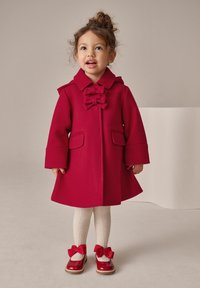 Rote Wolljacke mit Schleifendetail, weiten Ärmeln und zwei Vordertaschen. Kombiniert mit cremefarbenen, strukturierten Strumpfhosen und roten Schuhen mit Schleifen.